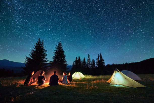 istock Group of hikers resting around campfire, surrounded by illuminated tents under stunning starry sky. 2202433232