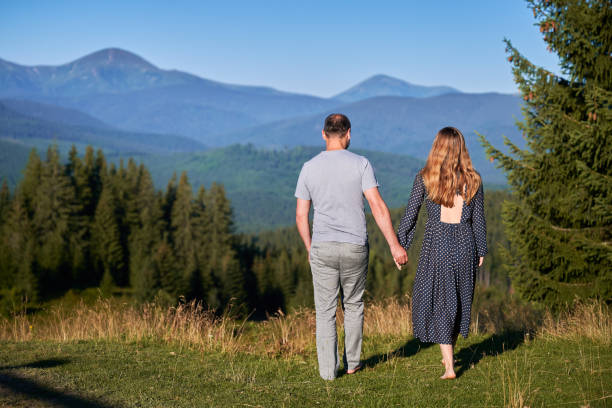 Couple holds hands, walk barefoot through meadow, facing stunning mountain view. Lush green forest and distant mountains under clear blue sky create serene and romantic atmosphere.