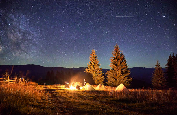 Blurred silhouettes of hikers resting around campfire, surrounded by illuminated tents under stunning starry sky. Milky Way stretches above, creating magical atmosphere in mountain wilderness.