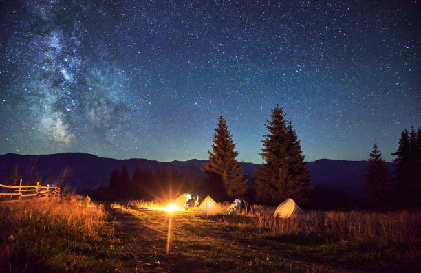 istock Group of hikers resting around campfire, surrounded by illuminated tents under stunning starry sky. 2202433272
