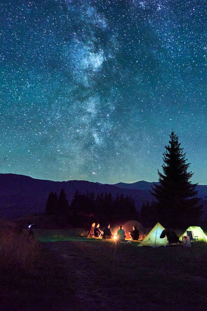 istock Group of hikers resting around campfire, surrounded by illuminated tents under stunning starry sky. 2202433281
