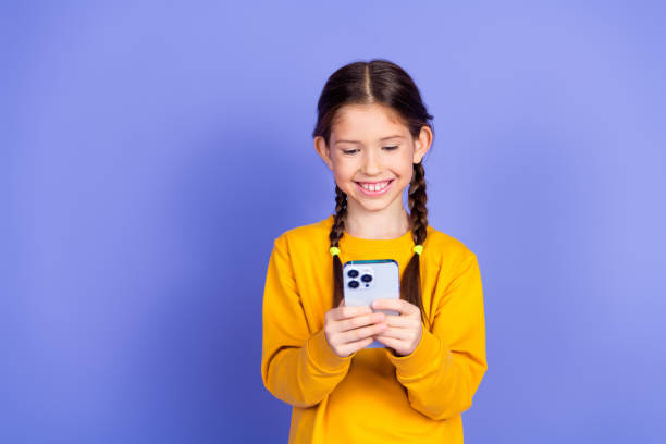 istock Smiling young girl using a smartphone against a vibrant violet wall, looking cheerful and trendy in orange sweatshirt 2202435471