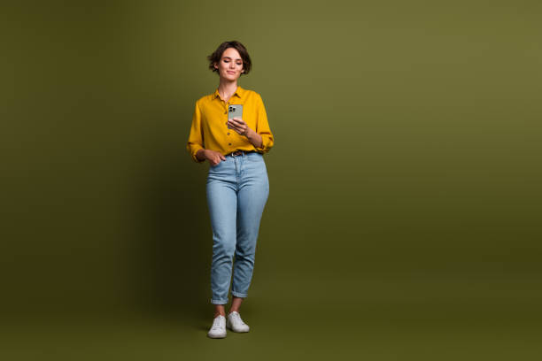 istock Smiling Young Woman in a Yellow Shirt Checking Her Smartphone While Standing on a Plain Olive Green Background 2202437909
