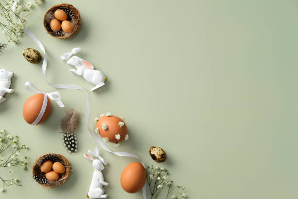 istock Happy Easter concept. Flat lay composition with decorated eggs, ceramic bunnies, bird nests, feathers, and white flowers on a light green background 2202442503