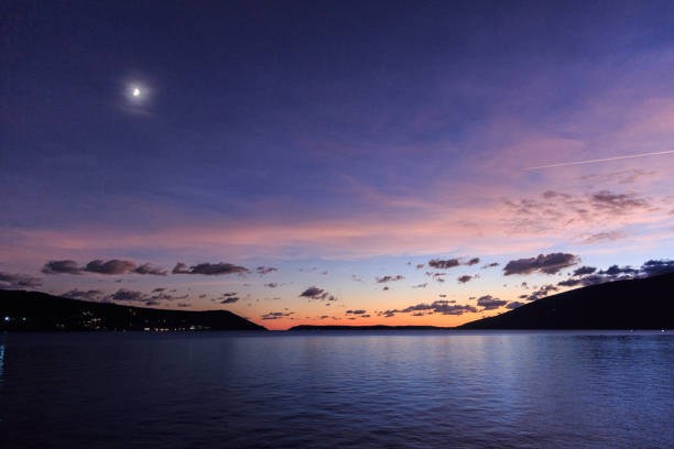 This image captures a breathtaking sunset over a tranquil body of water. The sky is ablaze with a stunning array of colors - deep purples, vibrant pinks, and warm oranges that dance across the clouds. A crescent moon hangs delicately in the upper left corner, adding to the serene and ethereal atmosphere.\n\nThe water below reflects the vibrant hues of the sky, creating a mirror-like effect that doubles the visual impact. In the distance, the silhouettes of mountains rise up, adding depth and dimension to the scene. \n\nThe overall feeling is one of profound peace and natural beauty. The stillness of the water, the softness of the light, and the vastness of the sky all come together to create a truly breathtaking landscape. This is a moment of pure tranquility, a chance to pause and appreciate the majesty of the natural world.