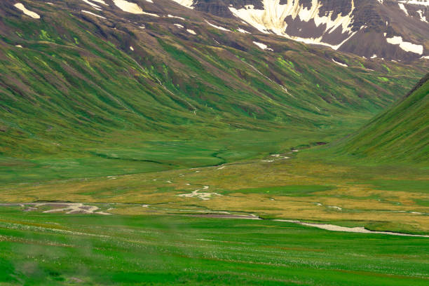 A lush green valley with a winding river, surrounded by towering mountains with patches of snow.
