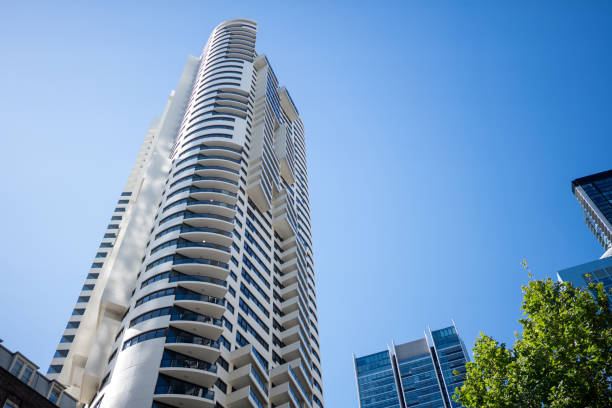 A low-angle view of a contemporary high-rise residential building with curved balconies and a sleek design. The skyscraper stands tall against a clear blue sky, with surrounding office towers and green foliage adding to the urban landscape.