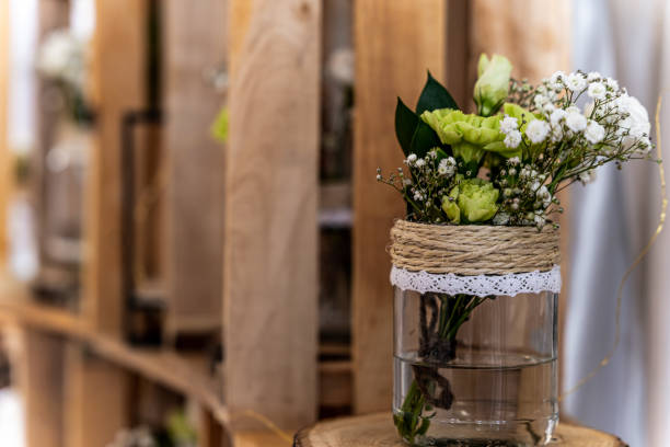 A wedding floral arrangement with green and white flowers in a glass jar wrapped with rustic rope and lace, placed on a wooden shelf