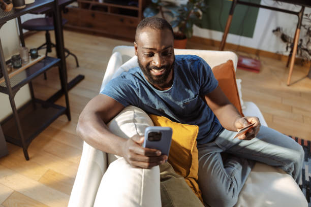 istock Man shopping online from home using smartphone on a cozy white couch 2202458309
