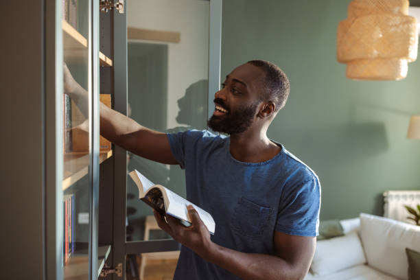 istock Man selecting a book from shelf holding a novel in a cozy home 2202458310