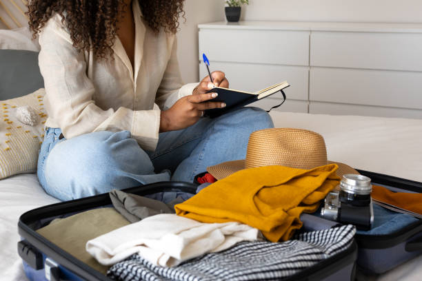 istock Young woman writing in notebook while packing suitcase for travel 2202462490