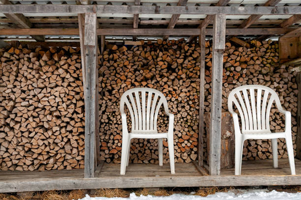 Two white plastic chairs sit in front of neatly stacked firewood in a wooden storage structure, surrounded by a snowy landscape.
