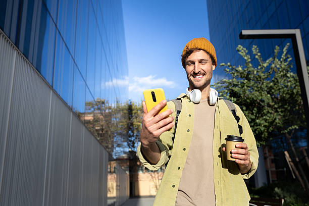 istock Smiling young man using smartphone and holding coffee walking in city 2202469353