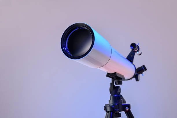 Detail of the main lens of a white telescope with eyepiece and finder scope mounted on black tripod isolated on blue and pink gradient background. Front view.