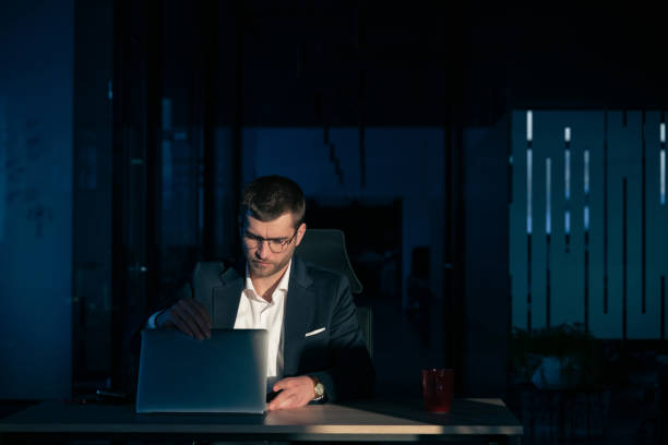istock Business executive in a dark office closing his laptop, illuminated by a single light source. Concept of finishing work, exhaustion, overtime, corporate routine, or end of a long workday 2202480196