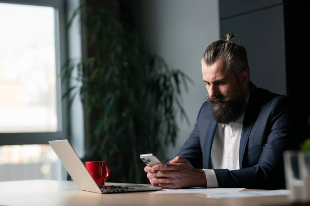 istock Businessman in a modern office, wearing a suit, looking at his smartphone while sitting at a desk with a laptop and a red coffee mug. Concept of digital communication and modern work environment 2202481070