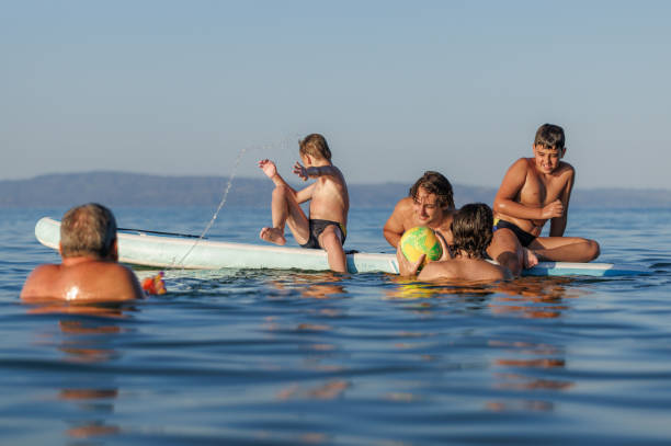 Laughter filling the air as two kids trying to maintain their footing on a floating surfboard, engaging in a friendly competition, their father joining in the challenge in the water
