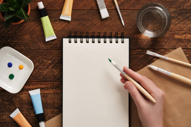 istock An artist's hand holds a brush over a blank sketchbook. A place for your text or image. 2202486305