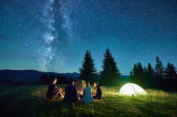 istock Group of hikers resting around campfire, surrounded by illuminated tents under stunning starry sky. 2202492656