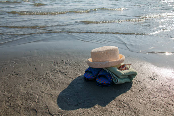 Beach accessories are spread out on the sand, featuring striped blue flip-flops, a rolled-up towel, and a straw hat. These items set the scene for a perfect day of sun and relaxation at the beach.