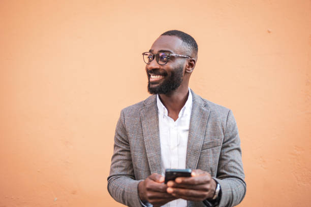 istock Confident businessman smiling while holding smartphone against peach wall 2202516827