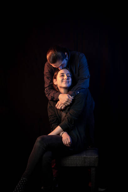 A loving couple shares a warm embrace in a professional studio portrait. The man kisses the woman's head while she smiles with contentment, expressing deep affection.