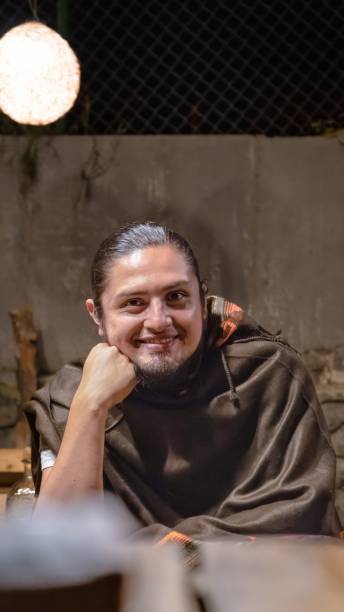 Young man with a confident expression wearing a traditional poncho, sitting in a rustic outdoor setting at night. Warm lighting and a cozy atmosphere