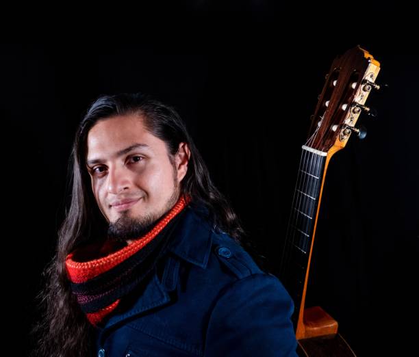 Portrait of a smiling musician with long hair and a beard, wearing a red scarf and navy coat, posing beside a classical guitar against a dark background.