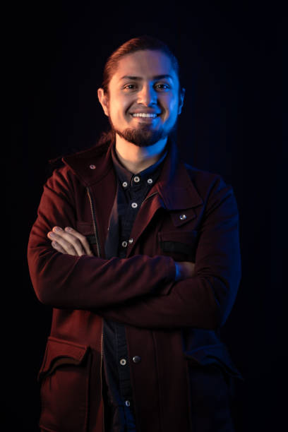 A confident man with a warm smile and crossed arms poses under moody blue and warm lighting, creating an artistic and stylish portrait against a dark background.