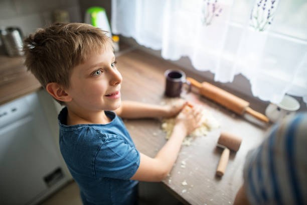 Young boy in a blue T-shirt is focused on his dough, looking with a smile at his sistert. The bright kitchen is filled with natural light coming through a window with sheer curtains.\nShot with Nikon D810