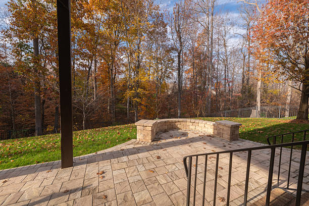 A backyard patio with stone pavers and a curved stone wall, framed by black metal railings. Fallen leaves and autumn foliage create a warm, seasonal atmosphere.
