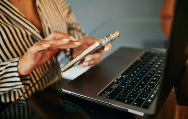 Close-up of a woman's hands using a mobile phone while working on a laptop in a modern digital home setting. The scene highlights efficient multitasking and the flexibility of remote work