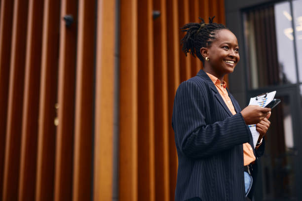 istock Smiling black businesswoman using smartphone and holding documents outside modern office building 2202543393