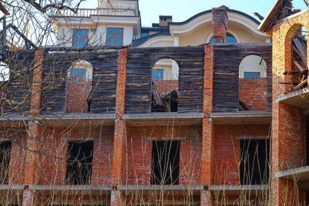 istock An unfinished brick house with arched windows and wooden elements. In the background, another house with a white facade can be seen. In the foreground, bare tree branches are visible. 2202545302
