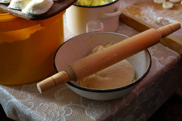A wooden rolling pin rests in a bowl of dough, on a table with various kitchen utensils.