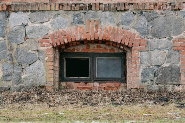 Decaying Wall with Cracked Bricks and a Broken Glass Window. Mysterious Basement Window in an Aged Stone Structure