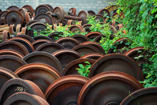 A large pile of rusty metal wheels, possibly from trains or industrial machinery, sits outdoors.  Vegetation, including green leaves and small plants, grows between and around the wheels, suggesting a state of disuse or abandonment.