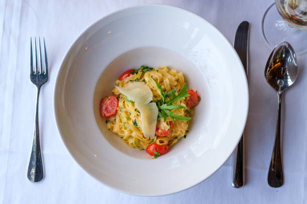 A delectable plate of pasta, featuring a creamy sauce, vibrant cherry tomatoes, fresh herbs, and a delicate parmesan shaving. The elegant presentation is set against a pristine white tablecloth.