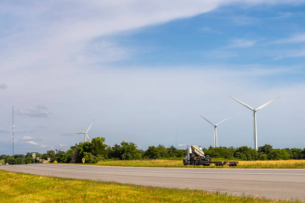 Three massive white wind turbines with slowly rotating blades dominate the skyline of a flat grassland landscape in Oklahoma. A concrete World War I Veterans Memorial highway cuts through the foreground, with a small truck visible in the distance. Lush green vegetation lines the roadside, while a partly cloudy sky stretches overhead