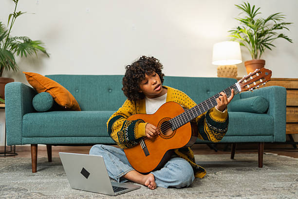 istock African American woman learning guitar watching tutorial on laptop at home. Black girl playing guitar singing song learning online music lessons, Artistic woman playing acoustic guitar learning chords 2202586891