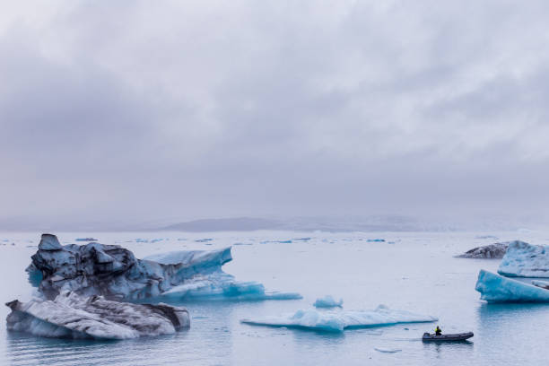 istock A serene yet captivating view of icebergs floating gracefully in a tranquil lagoon, with an adventurous individual navigating the water in a small boat under gloomy skies. 2202587432