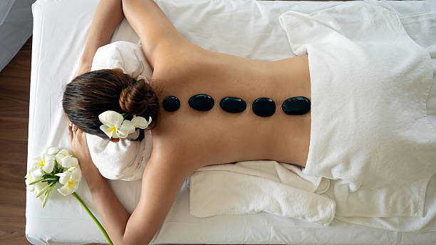 istock Young woman receiving hot stone therapy lying face down on massage table with black basalt stones aligned on her bare back, adorned with white flowers in hair 2202593335