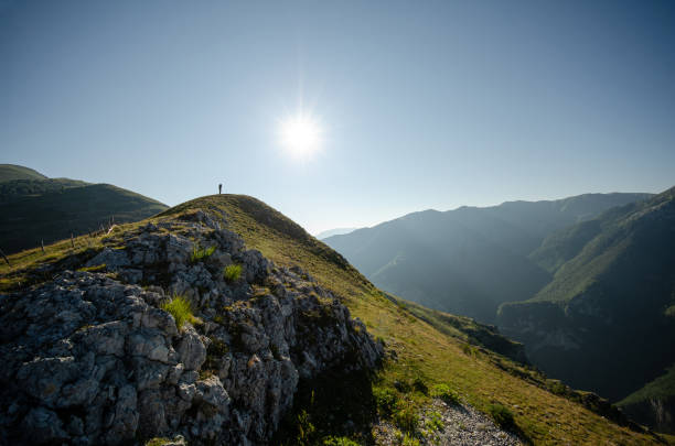 istock Lone figure atop a mountain ridge under a bright sun surrounded by rolling hills and distant peaks 2202599617