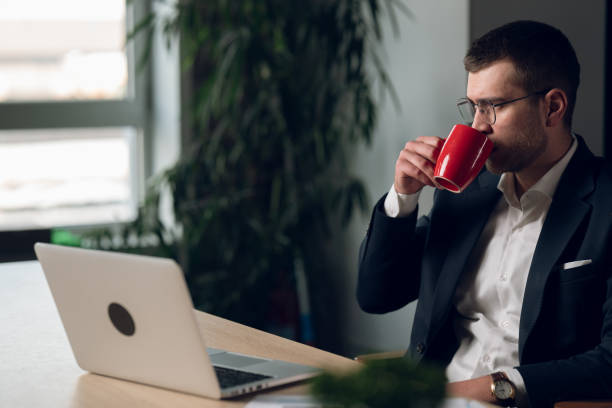 istock Professional man in glasses, wearing a suit, sipping coffee while working late at a desk. Concept of focus, productivity, and modern office lifestyle 2202624908