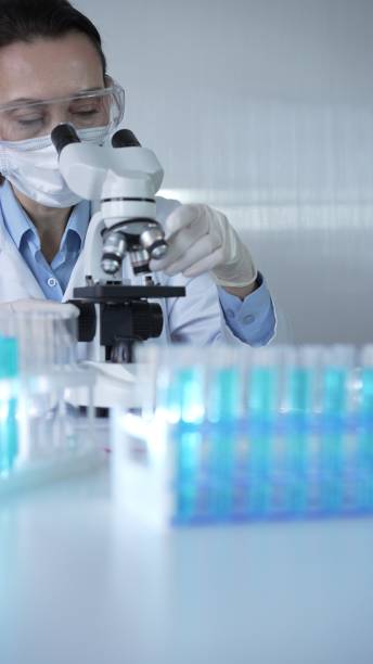 istock Woman scientist wearing lab coat, white protective gloves and glasses, is using microscope in modern laboratory setup, portrait view. Low key lighting photo. Science and Medicine concept 2202633384