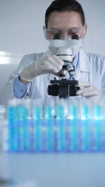 istock Woman scientist wearing lab coat, white protective gloves and glasses, is using microscope in modern laboratory setup, portrait view. Low key lighting photo. Science and Medicine concept 2202633901