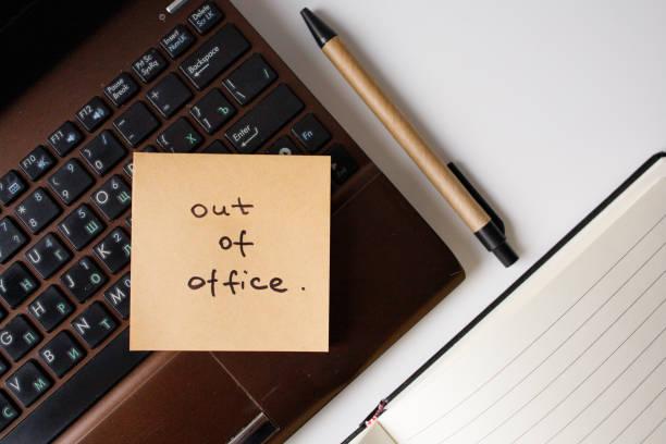 istock Sticky note with handwritten phrase out of office on a laptop with pen and notebook top view 2202646677