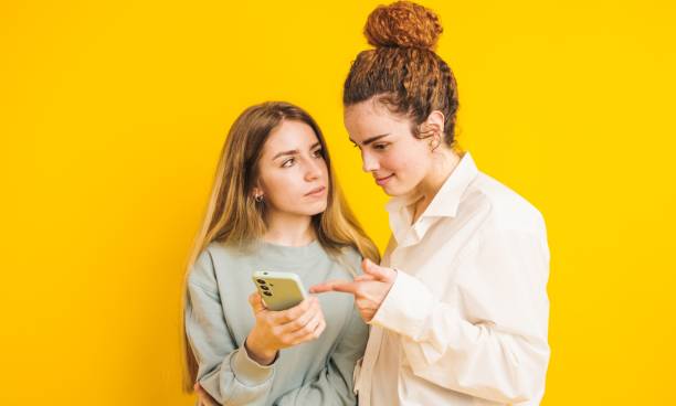 istock Two young women sharing a smartphone on yellow background 2202648108