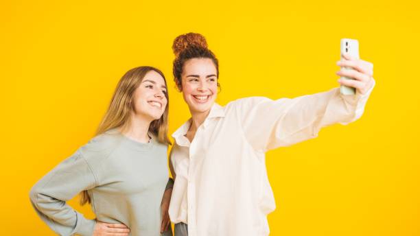 istock Two young women taking a selfie with smartphone on yellow background 2202648154