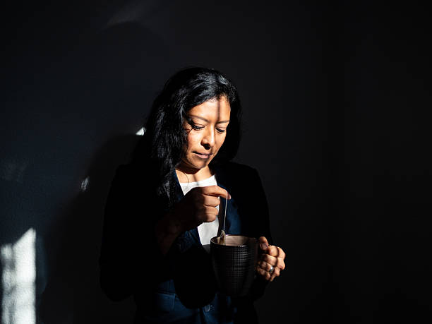 A Mexican woman in her thirties gently stirs a drink while standing in a dimly lit space. Soft shadows play across her face, enhancing the calm atmosphere of the moment.